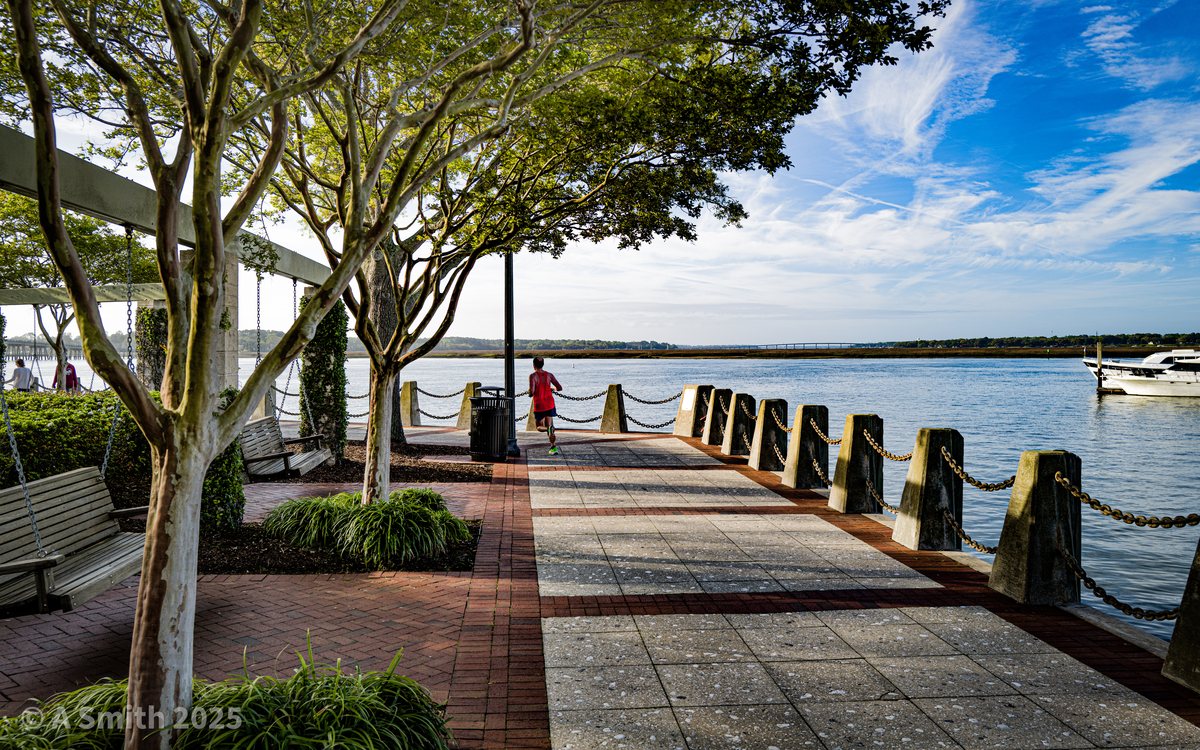 Shade on the riverwalk. The pergola stretch — benches, swings and shade facing the river.