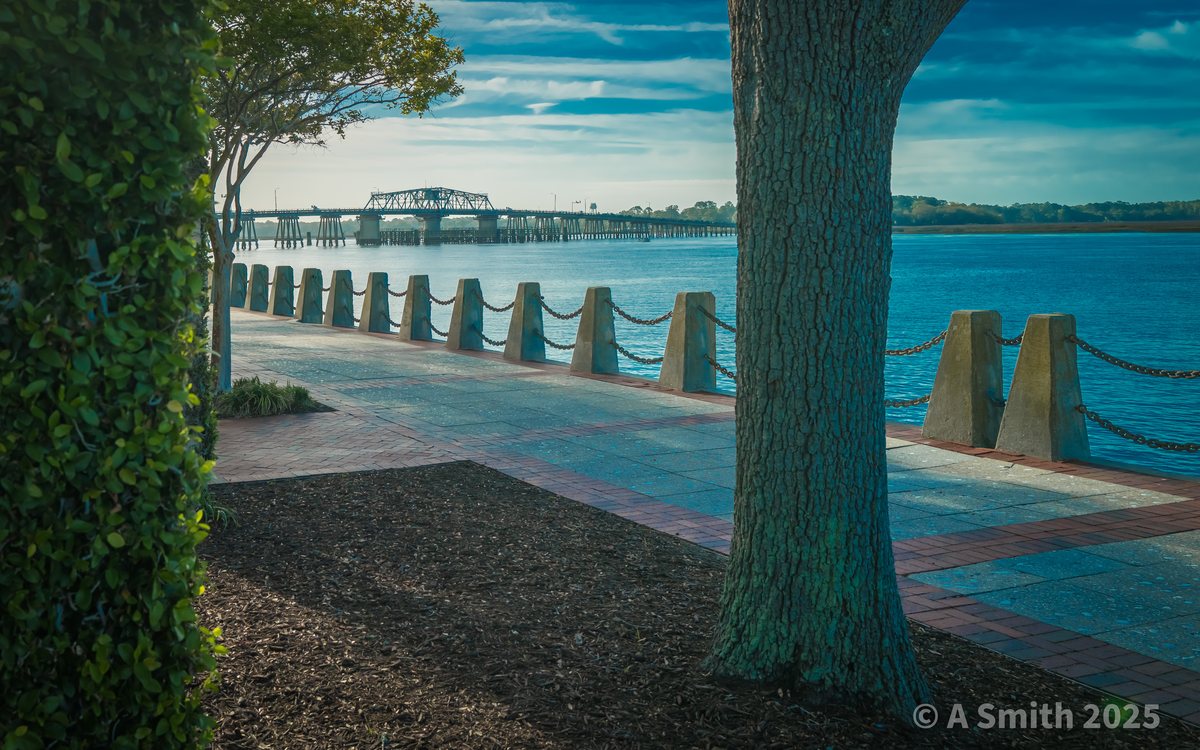 The promenade, teal and brick. The riverwalk running west from the park, the swing bridge low on the horizon.