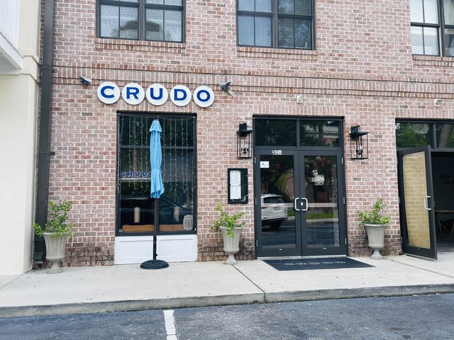 CRUDO at The Promenade in Bluffton — five circular letter signs on the brick wall, black doors and urns on the sidewalk