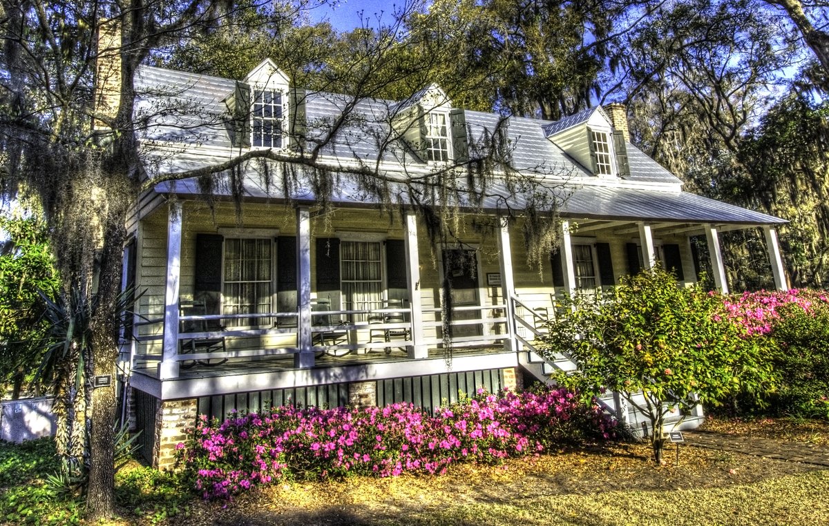 The Heyward House in Old Town Bluffton in full azalea bloom — pink flowers under the porch, rocking chairs, moss-draped oaks