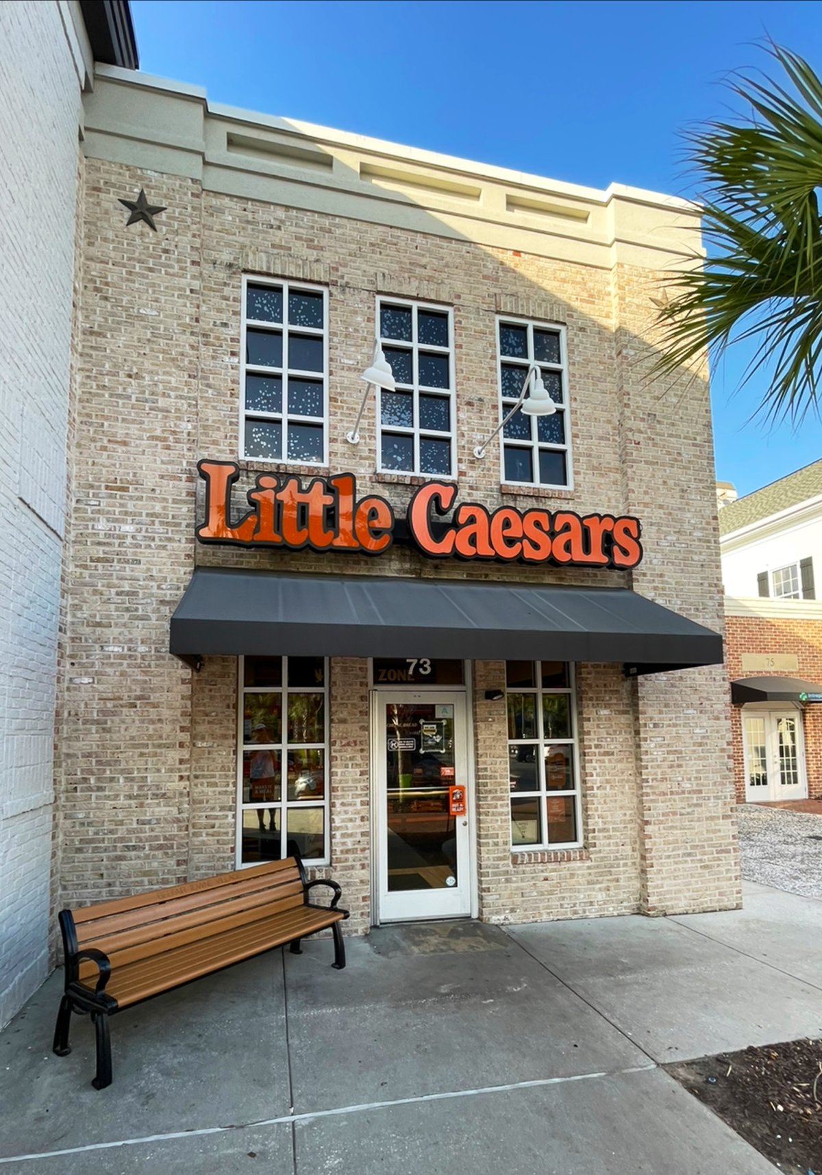 Little Caesars in Belfair Towne Village, Bluffton — orange letters on a two-story brick storefront, a bench out front