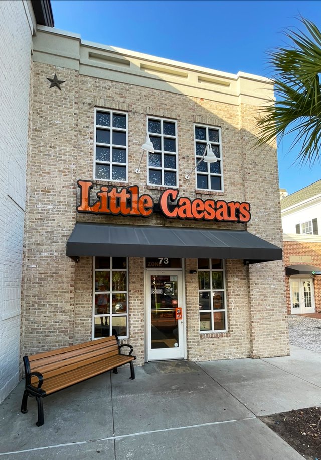 Little Caesars in Belfair Towne Village, Bluffton — orange letters on a two-story brick storefront, a bench out front