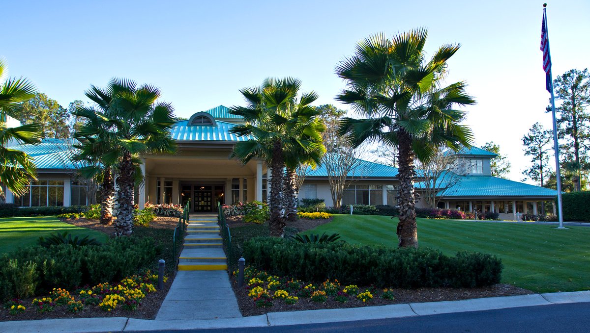 The clubhouse, wide. The clubhouse from the street — palms, the flag, and that teal roof.