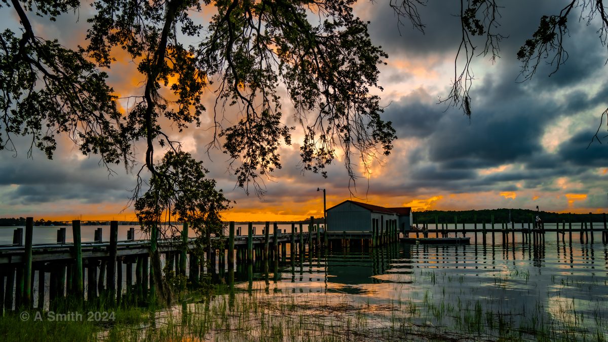 A dock and boathouse under oak branches at sunset, dramatic clouds over the water