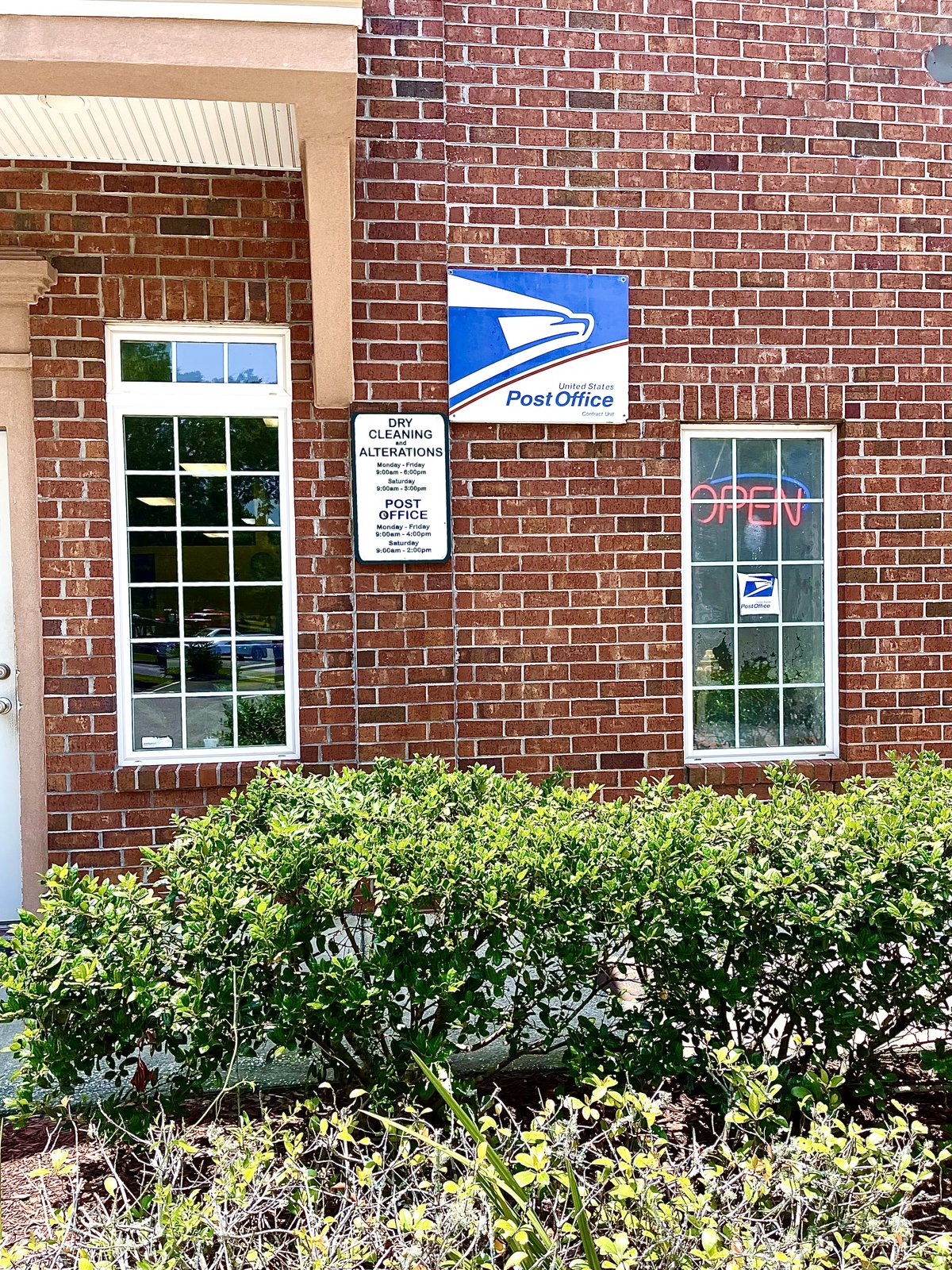 The post office counter in Okatie — the USPS eagle sign on a brick wall, a neon OPEN sign and the dry cleaning board beside it