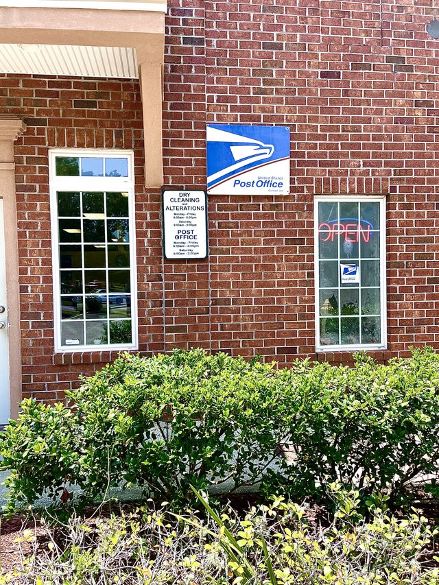 The post office counter in Okatie — the USPS eagle sign on a brick wall, a neon OPEN sign and the dry cleaning board beside it