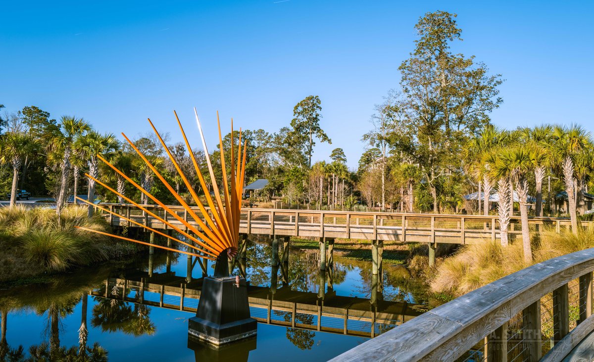 An orange sunburst sculpture rising out of a lagoon at Coligny, Hilton Head Island, a wooden boardwalk bridge behind it
