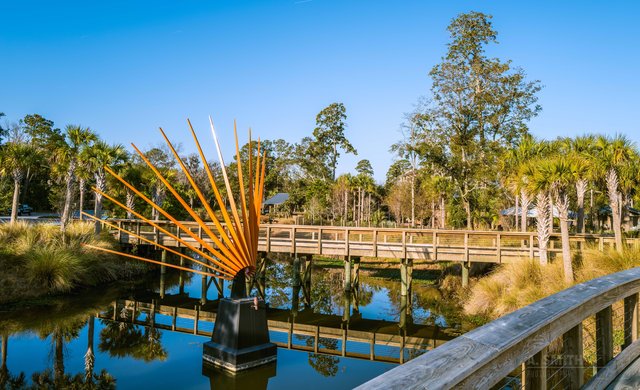 An orange sunburst sculpture rising out of a lagoon at Coligny, Hilton Head Island, a wooden boardwalk bridge behind it