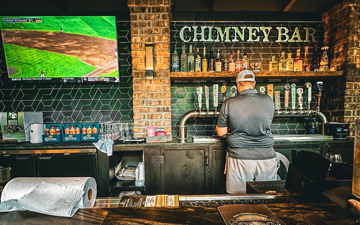 The Chimney Bar inside The Smokehouse — green tile and brick, bottles and beer taps, a baseball game on the screen